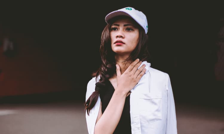 Brunette woman wearing a white hat, white jacket, and red lipstick puts her hand anxiously on her neck