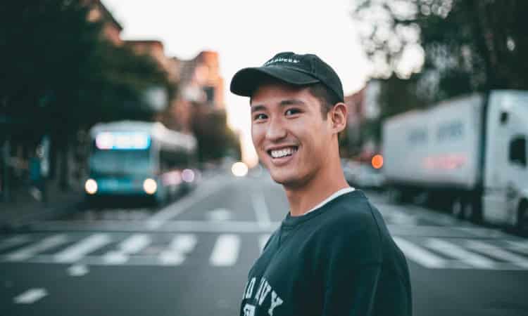 Brunette man smiles while crossing a street and wearing a ball cap and sweatshirt