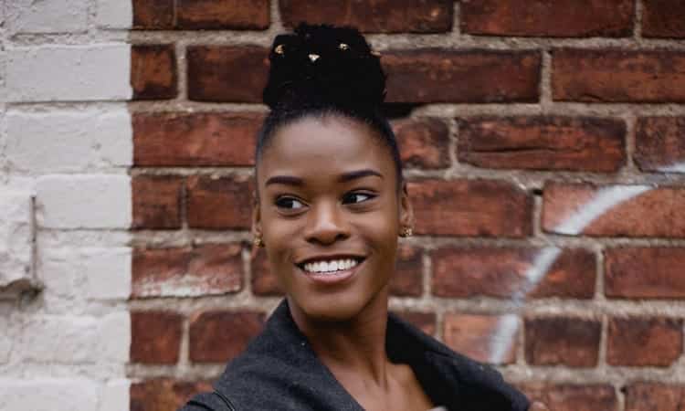 Dark-haired woman with her hair in a top bun smiles with white, living teeth in front of a red brick wall