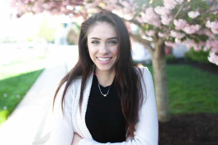 Dark-haired woman wearing a black shirt and sweater under a tree with pink blossoms smiles with straight, white teeth