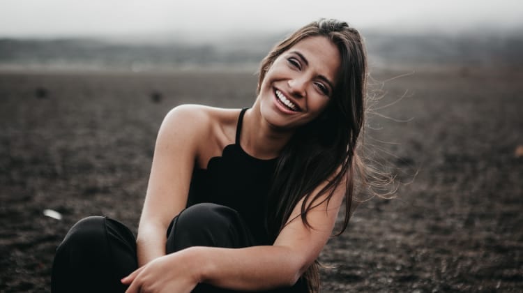 Brunette woman with porcelain dental veneers smiles while wearing a bank tank top and sitting on a dirt field