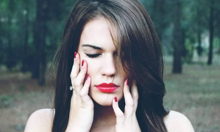 Brunette woman with red lipstick touches her fingers to the sides of her face due to sensitive teeth while standing in a forest